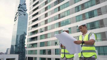 Engineer men are standing on a rooftop, looking at blueprints. They are wearing safety vests and hard hats. The building they are standing in front of is a large, modern skyscraper video