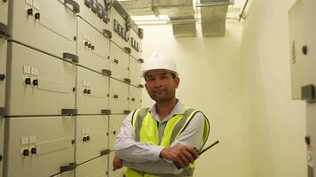 Engineer man in a yellow vest stands in front of a row of electrical boxes. He is wearing a hard hat and he is a worker in a factory video