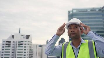 Engineer man in a yellow vest is standing on a rooftop. He is holding a hard hat and he is looking up at the sky. The scene suggests a construction site or a work area where safety gear is required video