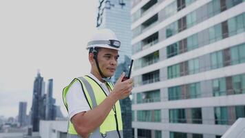 Engineer man in a yellow vest is holding a walkie talkie standing on a rooftop in front of a tall building video