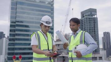 Two engineer men in safety vests are standing on a rooftop, looking at a tablet. Scene is serious and focused, as the men are likely discussing important information related to their work video