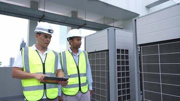 Two engineer men in yellow vests are standing next to a large air conditioner. One of them is holding a tablet video