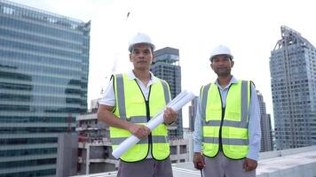 Engineer Two men in yellow vests stand on a rooftop in front of a building. They are wearing hard hats and holding blueprints video