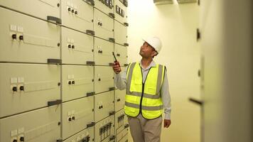 Engineer man in a yellow vest is looking at a row of white boxes. He is wearing a hard hat and is holding a walkie talkie video