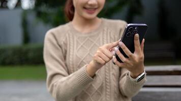 A close-up image of a happy Asian woman is using her smartphone on a bench in a garden. photo