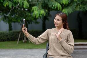 A positive Asian woman is taking a selfie or talking on a call on a bench in a park. photo