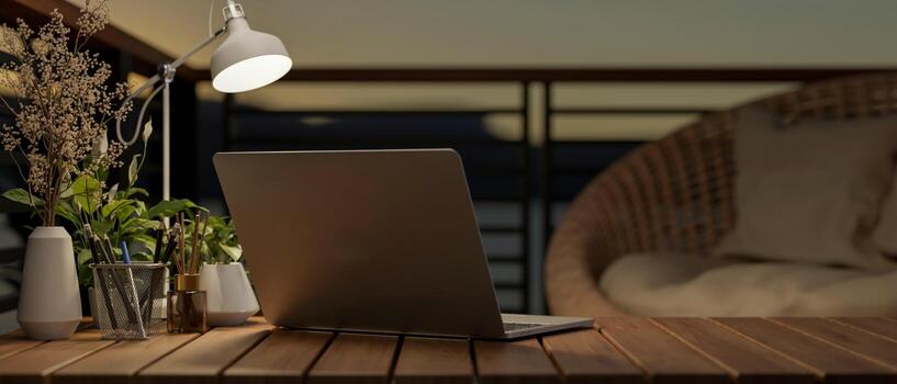 Back view image of a laptop computer on a wooden desk on a balcony at dusk, illuminated by a lamp. photo
