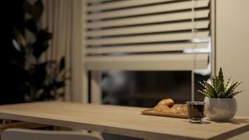 A wooden table against the window in a room features a coffee cup and a plate with a croissant. photo