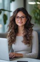 A cheerful woman with dark brown hair and glasses sits at her desk working on her laptop photo