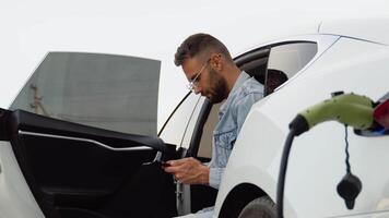 Bearded man with smartphone waiting for the finish of the battery charging process of electric car video