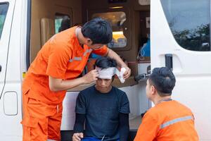 A man with a bandaged head is being helped by two other men in orange. The scene is serious and focused on the injured man photo