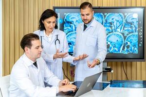 Team of three doctors discusses the results of an MRI scan of the patient's head in the consulting room of modern hospital, physician discussing about treatment for patient looking in laptop photo