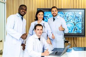 Group of medical practitioners in lab coats gesturing thumbs up while sitting and standing with smiling on camera. Four colleagues discusses the results of an MRI scan of the patient's head photo