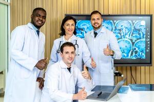 Group of medical practitioners in lab coats gesturing thumbs up while sitting and standing with smiling on camera. Four colleagues discusses the results of an MRI scan of the patient's head photo