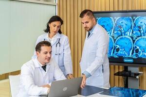 Team of three doctors discusses the results of an MRI scan of the patient's head in the consulting room of modern hospital, physician discussing about treatment for patient looking in laptop photo