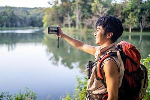 Young asian attractive male tourists takes a photo of a beautiful autumn nature with a view of a lake and a park with his phone and selfie. Walking in the park alone, admiring the beauty nature
