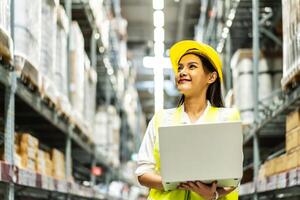 Smiling asian woman warehouse worker doing stocktaking of products management in cardboard box on shelves in warehouse using laptop computer, logistic and business export photo