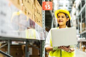 Smiling asian woman warehouse worker doing stocktaking of products management in cardboard box on shelves in warehouse using laptop computer, logistic and business export photo