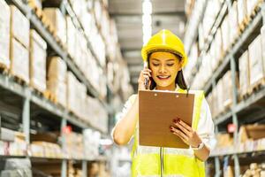 Asian woman in helmet and uniform talks on mobile phone discuss amount of stock product inventory on shelf at distribution warehouse factory. logistic business shipping and delivery service photo