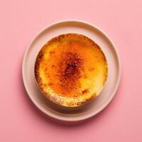 Close-up of a creamy, golden-brown creme brulee with a crispy, caramelized sugar topping on a white plate against a pink background. photo