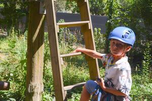 View of high ropes course, kids of climbing in amusement acitivity rope park, passing obstacles and zip line on heights, children teenagers in equipment gear between the trees on heights, summer day photo