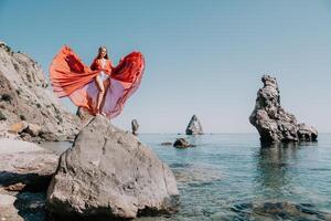 Woman travel sea. Young Happy woman in a long red dress posing on a beach near the sea on background of volcanic rocks, like in Iceland, sharing travel adventure journey photo