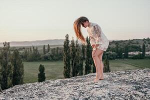 Happy woman in white boho dress on sunset in mountains. Romantic woman with long hair standing with her back on the sunset in nature in summer with open hands. Silhouette. Nature. Sunset. photo
