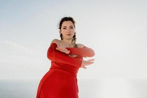 Closeup portrait of sensual young brunette woman in red dress, happily dancing outdoors isolated on blurry sea background with natural bokeh in soft warm sunset backlight. Selective focus. photo