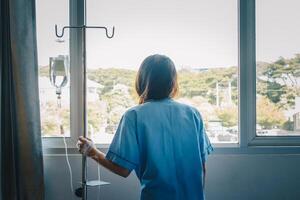 Back view of sickness patient woman standing nearly window in hospital ward, looking to outside and hope her health will be better. photo