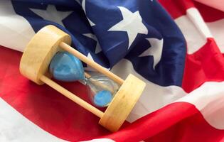 Hourglass on table with USA flag in the background. Sand glass on old wooden table with American flag in the background, vertical image. photo