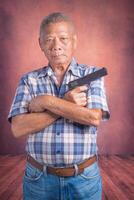 Portrait of an elderly Asian man holding a short gun and looking at the camera while standing with a brown background. Space for text. Concept of aged people and sport photo