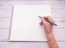 Hands of a senior woman writing on notebook with a pen on wooden table background. Space for text. Top view photo