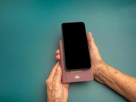 Hands of a senior woman holding a passport and smartphone on a green background. Space for text. Close-up photo. Aged people, communication and travel concept photo