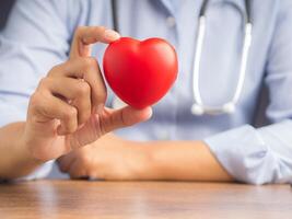 Hand of a doctor showing a red heart shape while sitting in the hospital or clinic. Close-up photo. Space for text. Medical and encouragement concep photo