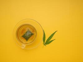 The top view of hemp tea in a teacup and marijuana leaves isolated on a yellow background. Cannabis herbal tea. Flat lay. Selective focus with copy space for text. Relaxation concept photo