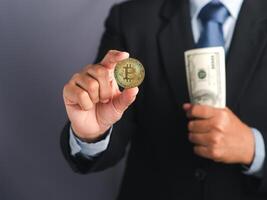 Hand of a businessman showing a bitcoin golden coin and holding US dollars banknotes while standing with a gray background. Close-up photo. Virtual cryptocurrency and future money concept photo