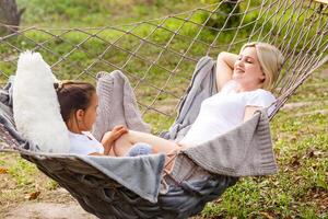 Wide view of a young mother and daughter relaxing together and smiling sitting in a hammock, hugging and lounging during a sunny summer day in a holiday home garden with grass and trees, lifestyle photo