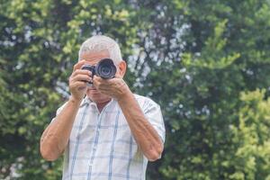 An elderly Asian man is taking photographs with a digital camera while standing in a garden. Aged people photographer and relaxation concept photo