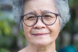 Portrait of an elderly woman with short gray hair, wearing glasses, smiling, and looking at the camera while standing in a garden. Aged people and relaxation concept photo