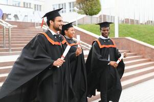 Group of happy smiling multicultural people in graduation gowns and caps with diplomas in hands graduate university outdoors in campus looking at camera. Graduation from college, university concept. photo