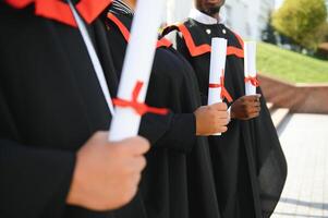 grupo de contento sonriente multicultural personas en graduación vestidos y tapas con diplomas en manos graduado Universidad al aire libre en instalaciones mirando a cámara. graduación desde colega, Universidad concepto. foto