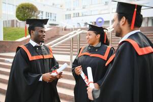 Group of happy smiling multicultural people in graduation gowns and caps with diplomas in hands graduate university outdoors in campus looking at camera. Graduation from college, university concept. photo