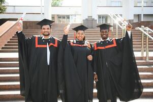 Group of happy smiling multicultural people in graduation gowns and caps with diplomas in hands graduate university outdoors in campus looking at camera. Graduation from college, university concept. photo
