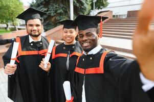 Group of happy smiling multicultural people in graduation gowns and caps with diplomas in hands graduate university outdoors in campus looking at camera. Graduation from college, university concept. photo