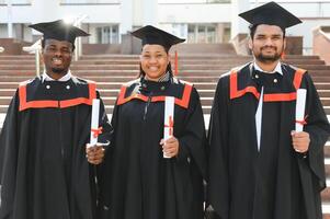 Group of happy smiling multicultural people in graduation gowns and caps with diplomas in hands graduate university outdoors in campus looking at camera. Graduation from college, university concept. photo