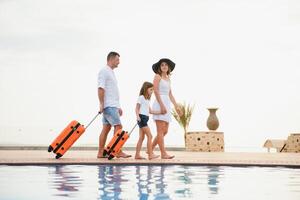 young family with suitcases walking to hotel building with beautiful swimming pool. traveling and relaxing time concept. photo