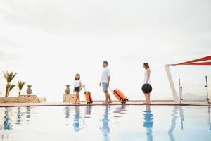 young family with suitcases walking to hotel building with beautiful swimming pool. traveling and relaxing time concept. photo