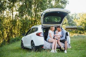 Pretty young married couple and their daughter are resting in the nature. The woman and girl are sitting on open car boot. The man is standing near them. They are smiling photo