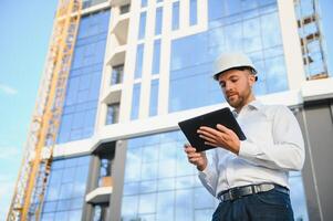 Engineer next to building cranes. Concept - large construction project. Architect directs the construction process. Drawings and tablet in the hands. photo