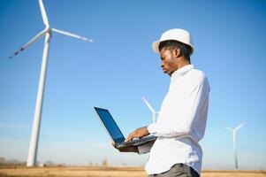 African engineer wearing white hard hat standing with digital tablet against wind turbine photo